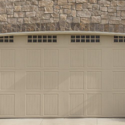 Beige sectional garage door with rectangular panels, set beneath a stone veneer wall on a suburban home.