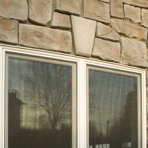 Double-pane window set in a rough stone wall, reflecting trees in the glass, with a triangular outdoor light fixture above.