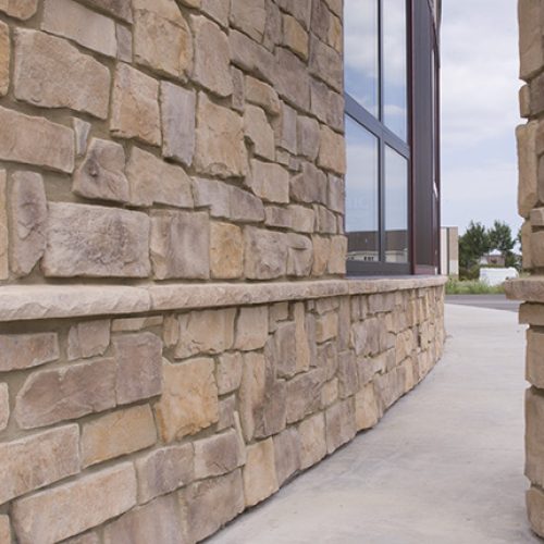 Exterior stone wall of a building with large beige-brown stones and a window reflected in the glass along a concrete sidewalk.