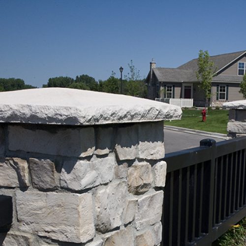 Stone pillar with a white capstone and black metal fence along a suburban street.