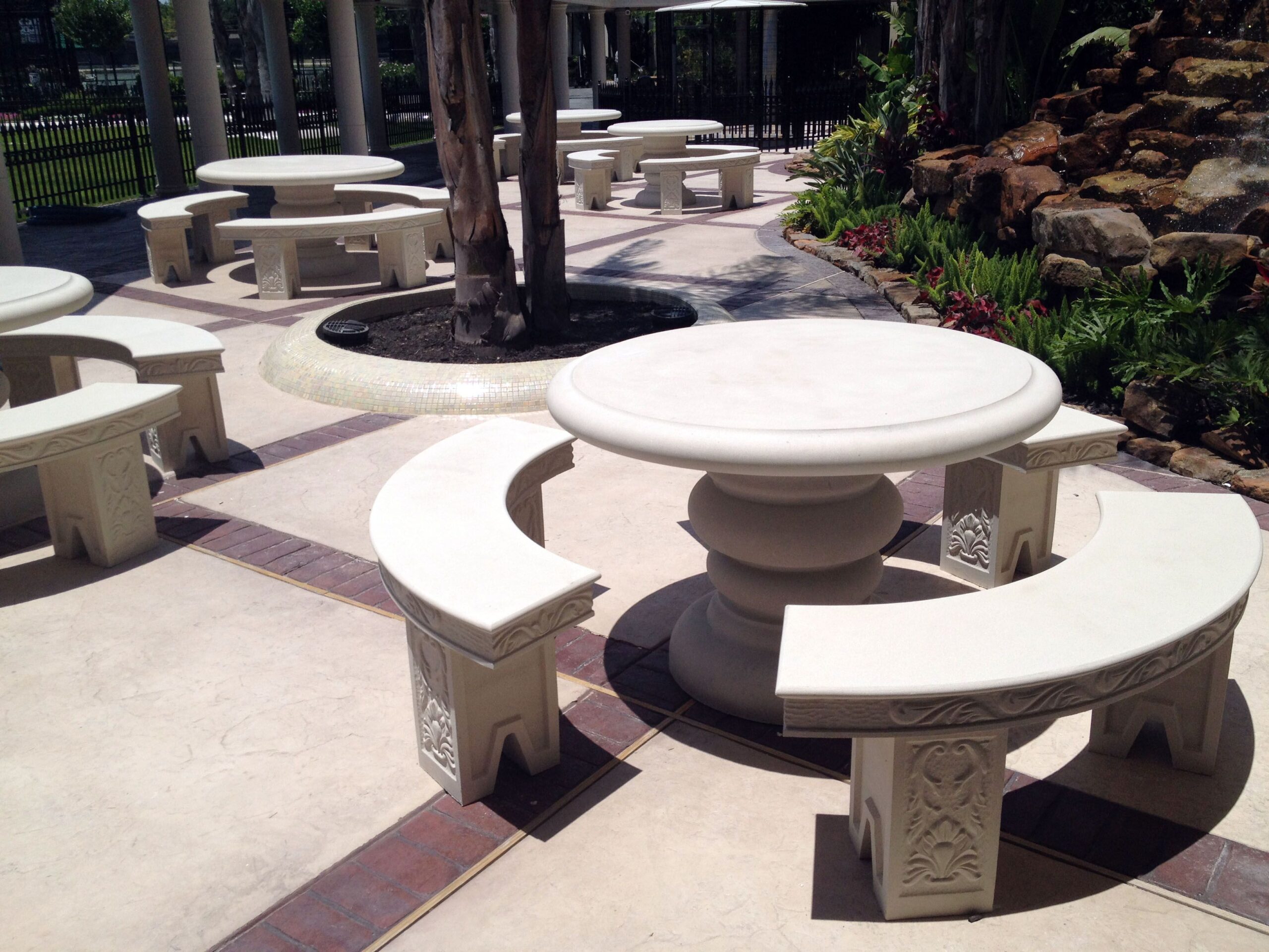 Outdoor courtyard with circular stone tables and benches around tree trunks, light stone paving, and a rock garden on the right.