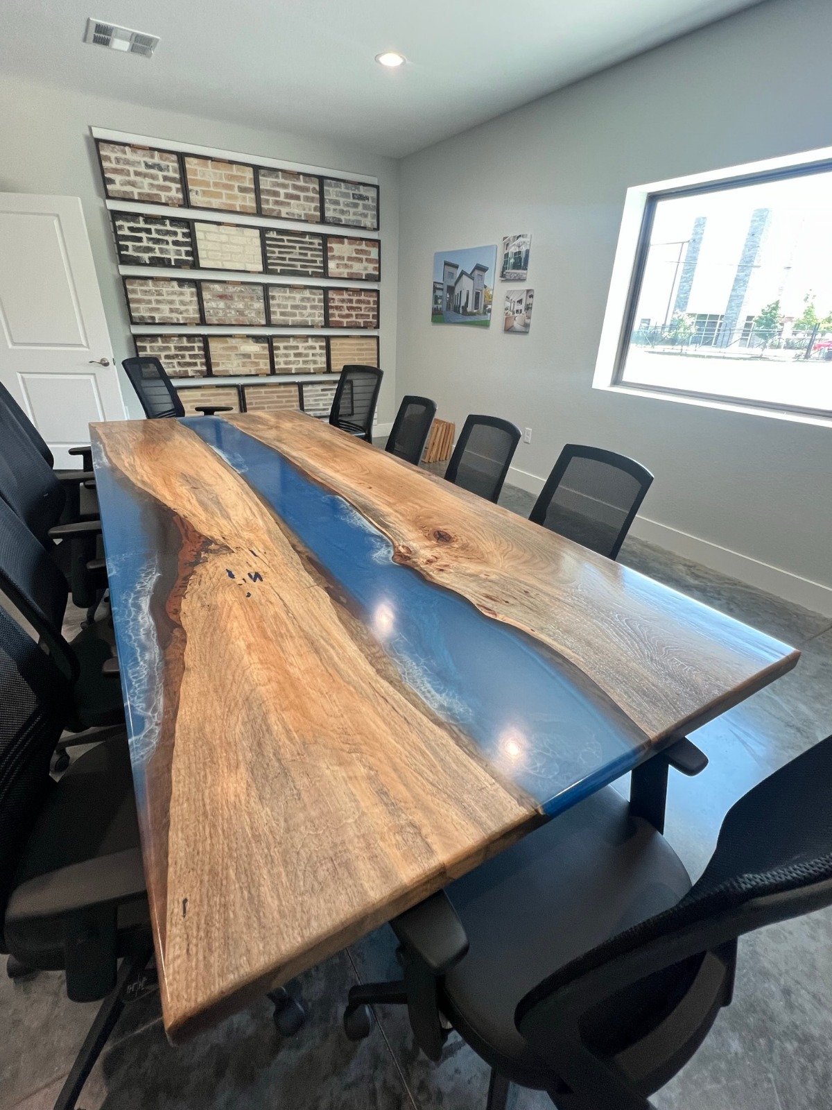 Long live-edge wooden conference table with a blue resin river, surrounded by black chairs in a bright meeting room.