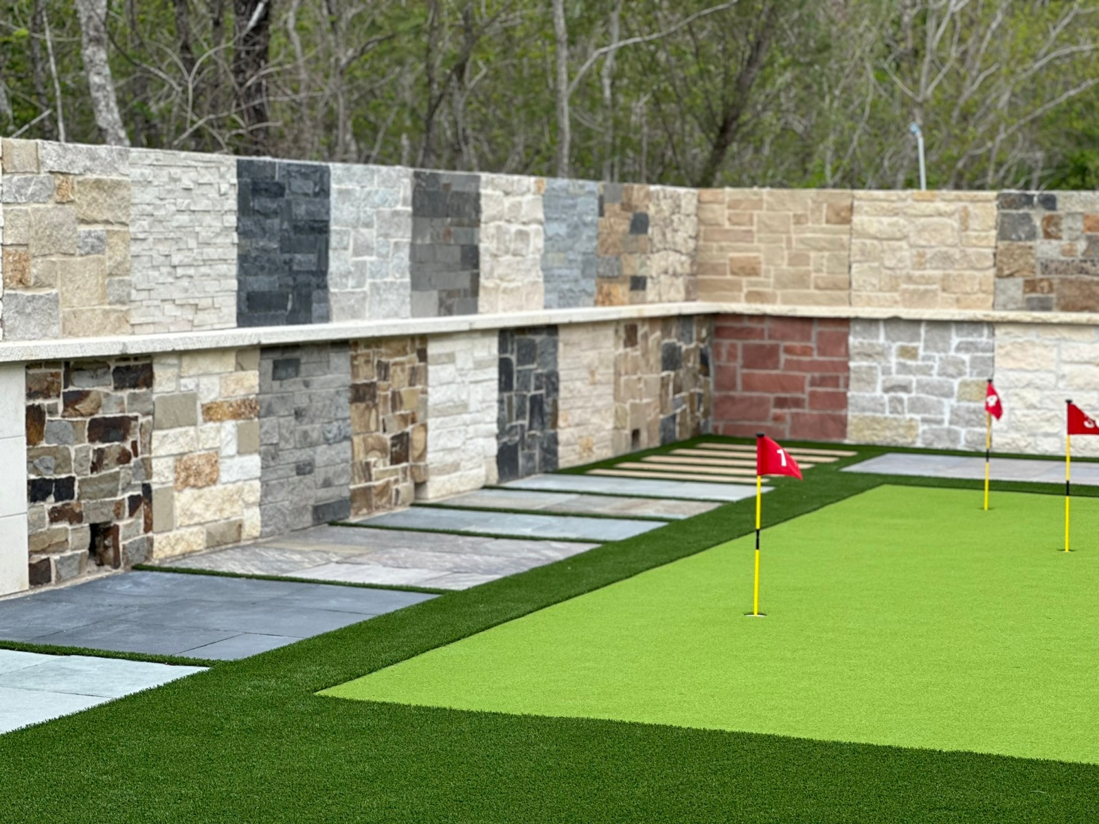 Outdoor mini-golf scene: bright green putting surface with red flags and a stone wall backdrop.