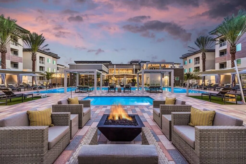 Resort-style pool deck at dusk with a central fire pit, wicker lounge chairs, and palm trees