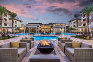 Resort-style pool deck at dusk with a central fire pit, wicker lounge chairs, and palm trees